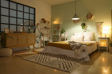 Interior of bedroom with bed, chest of drawers, shelving unit, bicycle and glowing lamps