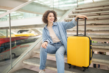 A cheerful woman with curly hair sits on the stairs of a busy terminal, resting beside her bright yellow suitcase. She enjoys a moment of relaxation amid the travel hustle and bustle.