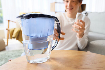 Young man with glass taking water filter jug from table at home, closeup
