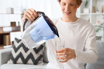 Young man pouring water from filter jug into glass on sofa at home, closeup