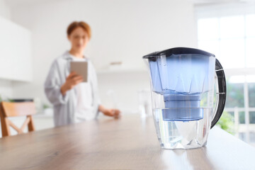 Water filter jug on table against man with tablet computer in kitchen, closeup