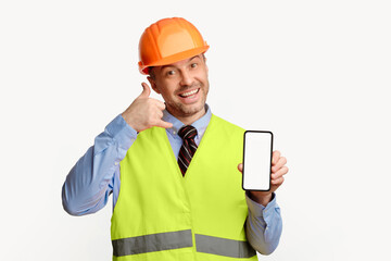Builder Man Showing Phone Blank Screen Gesturing To Call Him Posing Over White Studio Background. Building Services Concept. Mockup