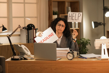 Young African-American businesswoman holding papers with words DEADLINE and HELP in office at night