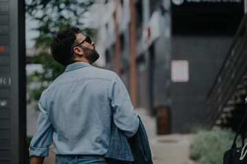 A businessman wearing sunglasses walks through an urban setting, holding a jacket and looking toward a building. The image captures a moment of casual, professional style in a cityscape.