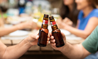 Two friends raise their beer bottles in a toast while enjoying a lively gathering outdoors. Other guests can be seen in the background, contributing to a cheerful atmosphere.