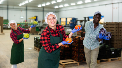 Two caucasian women and african-american man vegetable factory workers performing funny dance with vegetables.