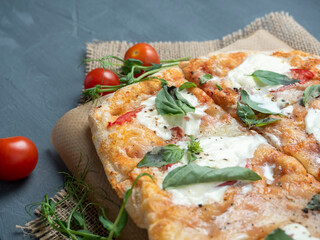 Close-up of delicious fresh crispy Roman pizza with cheese and herbs on a gray background. Fresh ripe tomatoes are nearby. Craft paper, linen napkin. Side view, selective focus