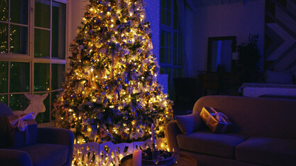 Decorated christmas tree with fairy lights is standing in a dark living room, illuminating wrapped gifts on armchairs