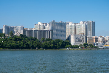 Modern high-rise residential buildings line the waterfront in Macao, China. A lush green park buffer stretches along the shoreline. Parque de Reservatório