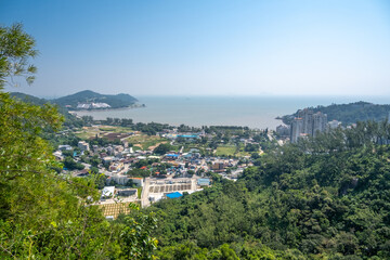 A scenic aerial view of Hac Sa Beach area in Macau, China with a mix of modern buildings, residential developments, and recreational facilities. Hills, and lush vegetation frame the coastline