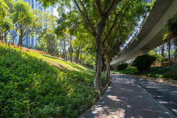 A pedestrian path winds through a lush, green park, with mature trees providing shade, manicured...