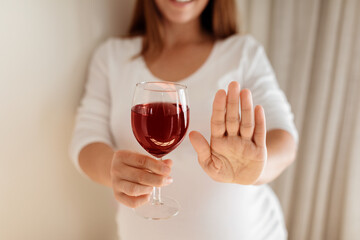 A pregnant woman is holding a glass of red wine while raising one hand in a stop gesture. She...