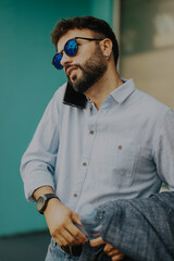 A stylish man wearing sunglasses and a casual shirt converses on his phone while standing outside. He embodies a relaxed, confident demeanor, showcasing modern fashion and a casual lifestyle.