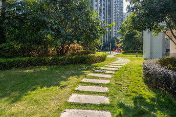 A winding stepping stone pathway leading through lush green grass, bordered by manicured hedges and trees, with a small playground in a park or communal garden within a residential apartment complex.