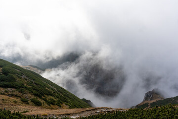 Clouds of mist rolling over green mountain slopes with a rocky outcrop partially visible. Concept of dramatic landscape, nature is beauty, and hiking through foggy wilderness. High quality photo