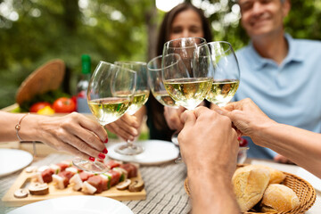 A group of friends toasting with glasses of white wine at a picnic table, surrounded by fresh food, lush greenery, and a festive atmosphere during a sunny day.