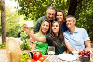 A group of five friends gathers around a table covered with fresh fruits and drinks. They smile joyfully as they take a selfie together in a vibrant, green park during a sunny day.