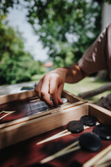 A close-up image of a hand playing a board game outdoors in a sunny garden. The scene captures a relaxed and strategic atmosphere amidst lush greenery.