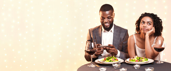 Bored young black woman at date in restaurant, afro man chatting on mobile phone