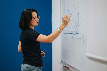 A young woman wearing glasses is explaining a diagram on a whiteboard in a classroom setting. She's...