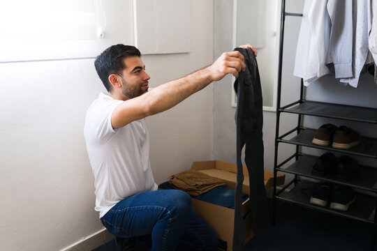 Young man holding a pair of pants while choosing an outfit from a cardboard box in his bedroom