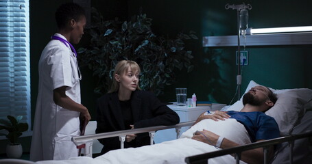 Woman sits next to an unconscious man in the hospital room as a nurse arrives.