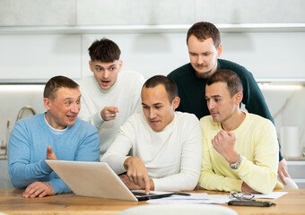 Fototapeta premium Group of men engaged in interested discussion with paperwork and laptop in cozy home kitchen during friendly meeting..