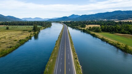 A scenic aerial view of a straight road bordered by a flowing river, surrounded by green fields and mountains under a blue sky.