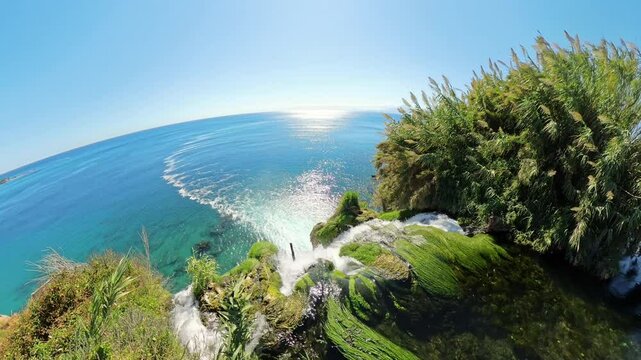 The camera captures the final moments as a river flows over the edge of a cliff into the sea, with lush greenery surrounding the waterfall and the vast ocean stretching into the horizon under a clear 
