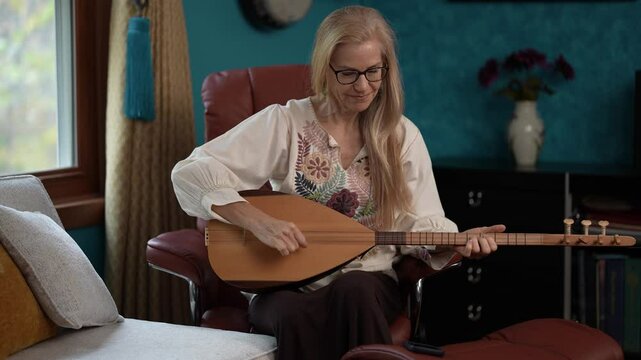 A woman with long blonde hair sits in a brown leather chair playing a saz instrument. She is wearing a white shirt with floral embroidery and black pants. 