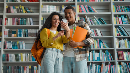 Two students classmates African American man Indian Latino Arabian woman mobile phone smartphone library smile pleasure laugh communication internet web girl guy education university learning study
