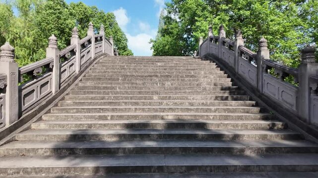 Walking up on a traditional Chinese stone arch bridge with curved railings and ornate carvings, leads up a set of stairs to a higher level in public park or garden.