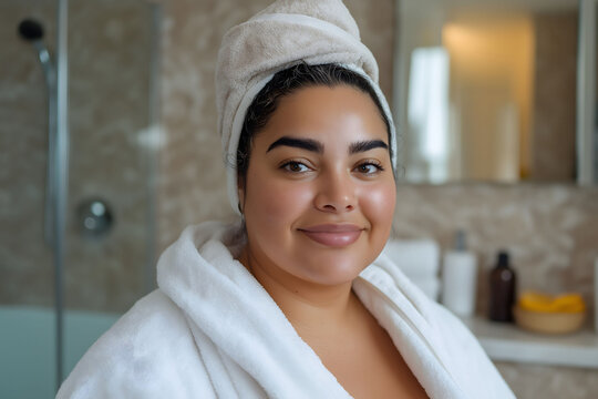 Curvy woman is wearing a bathrobe and a towel on her hair while smiling in her bathroom
