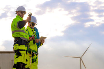 Two engineers wearing high visibility safety jackets and helmets measure wind speed with an...