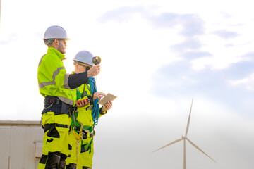 Two engineers wearing high visibility safety jackets and helmets measure wind speed with an anemometer and analyze data on a tablet at a renewable energy site. The cloudy sky adds depth to the scene.