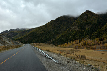 A straight asphalted road going up to high mountains with peaks in the clouds and a dense coniferous forest on a cloudy autumn day.