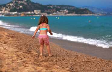 Fototapeta premium Beautiful fun happy kid girl walking in summer clothing on the yellow sand of the beach near blue sea and looking on water background. Happy