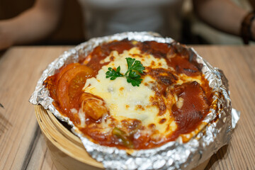 Cheese baked with tomato sauce at the base, covering rice and meat, served in a bamboo tray.