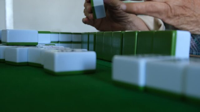 An elderly person's wrinkled hands, showing signs of age, hold a group of mahjong tiles and arrange them on the table. This traditional Chinese game requires skill and strategy.