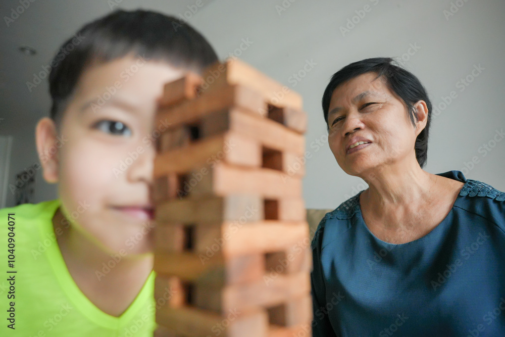 Wall mural Asian Young boy playing with wooden blocks while grandmother smiles in the background, joyful and focused expression, family bonding activity