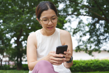 Asian Woman wearing eyeglasses sitting in a park and using a smartphone after workout exercise outdoor.