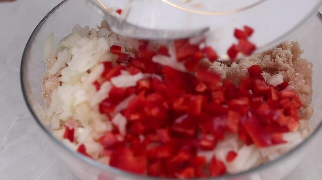 Close up of hands adding chopped vibrant red bell peppers and fresh onions to a clear glass bowl, containing minced chicken for patty burger preparation, capturing the culinary preparation