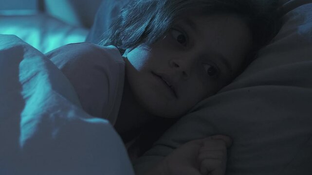 Night fear. Bedtime worry. Sleep disorder. Small girl woke up scared of bad dream at night lying in bed alone in dark home bedroom with blue moonlight.