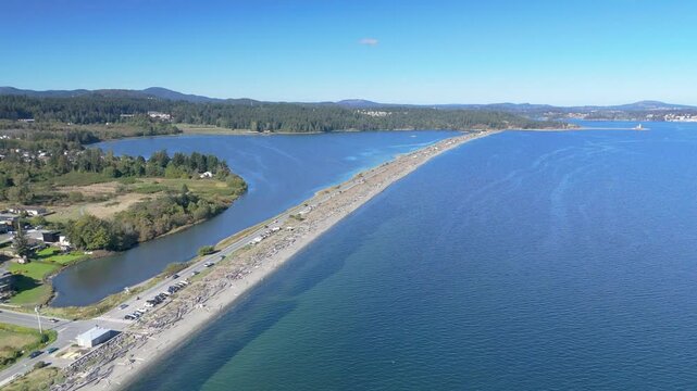Aerial view of a long, narrow strip of land extending into expansive blue waters, separating a calm bay from the open sea. Verdant greenery lines the shore, with scattered residential areas. 