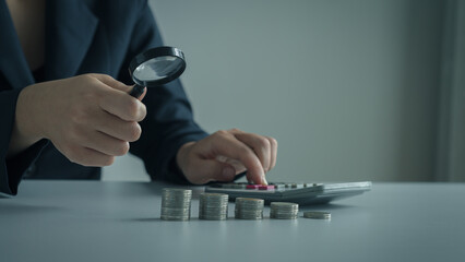 Fototapeta premium Businesswoman examining stacked coins with a magnifying glass while calculating finances, emphasizing financial analysis and precision.