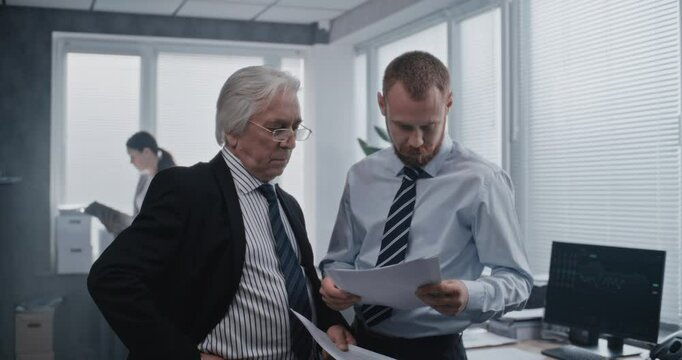 Two employees of international corporation with serious focused expression talking about business strategy and analyze financial reports on papers. Work in modern office of stock exchange company.