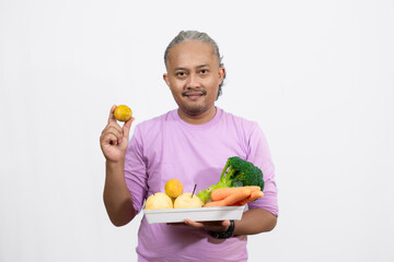 Obraz premium An Indonesian curly long-haired man holding a orange while carrying a tray of fruit and vegetables, healthy life concept 