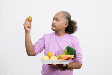 An Indonesian curly long-haired man holding a orange while carrying a tray of fruit and vegetables,...