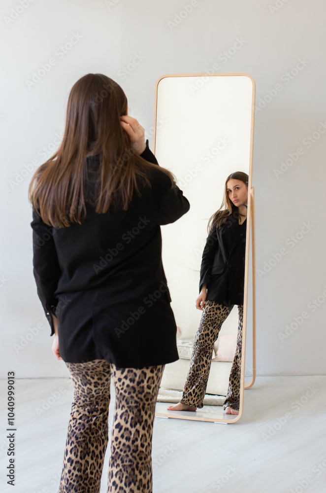 Poster back of young woman standing in front of mirror in black blazer and leopard print pants poses confid