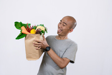 A bald, middle-aged Indonesian man wearing a grey t-shirt looks enthusiastic standing carrying a container filled with various kinds of vegetables and fruit, a healthy lifestyle concept.